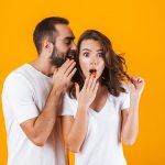 man telling a woman something in her ear, plain background.